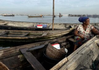 Eine Frau sitzt in einem Holzboot auf einem ruhigen Gewässer, umgeben von anderen Booten und Körben, mit entfernten Booten und einer Stadtlandschaft im Hintergrund.