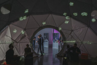 In einem überdachten Ausstellungsraum versammeln sich Menschen im Inneren und um eine geodätische Kuppelstruktur, auf deren Oberfläche grünes Licht projiziert wird.