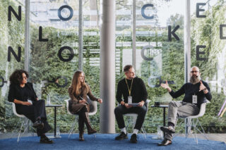 Vier Personen sitzen in einem modernen Raum auf Stühlen und diskutieren. Große Glasfenster im Hintergrund zeigen Pflanzen und Buchstaben. Jeder hat ein Schlüsselband und einen Notizblock dabei.