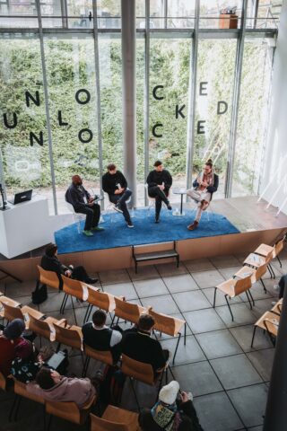 Ein vierköpfiges Gremium sitzt auf einer Bühne vor einem Publikum in einem modernen Hörsaal mit großen Fenstern und dem Wort „UNLOCKED“ an der Außenseite.