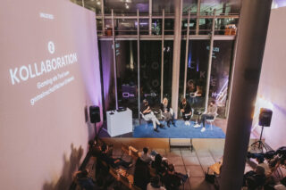 Bei einer Indoor-Veranstaltung sitzen vier Personen vor Publikum bei einer Podiumsdiskussion, an die Wand werden „Kollaboration“ und ein deutscher Text projiziert.