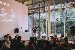 Eine Person steht auf der Bühne und hält einem Publikum in einem modernen Veranstaltungsraum mit Glaswänden eine Präsentation zum Thema Gamedesign.