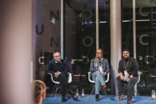 Drei Männer sitzen während einer Podiumsdiskussion auf Stühlen auf der Bühne. Sie wirken engagiert und haben jeweils eine Wasserflasche zu ihren Füßen. Im Vordergrund sind Zuschauer teilweise zu sehen.