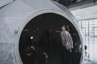 Eine Gruppe von Menschen sitzt und steht in einem weißen, kuppelförmigen Zelt mit einem großen runden Eingang in einem geräumigen Innenbereich.