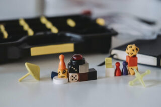 Auf einer weißen Fläche sind verschiedene bunte Brettspielsteine, Würfel und Spielfiguren angeordnet, im Hintergrund befinden sich eine schwarze Spielschachtel und ein Notizbuch.