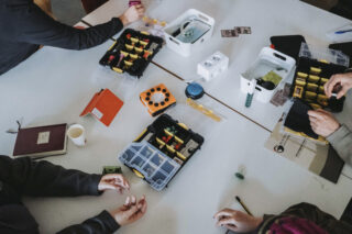 Menschen sitzen um einen Tisch herum und arbeiten mit elektronischen Bauteilen, Kabeln und kleinen Werkzeugkästen; auf dem Tisch liegen auch verschiedene Gegenstände und Notizbücher.