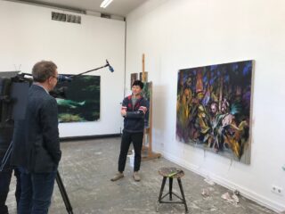 Ein Mann steht in einem Kunstatelier und wird vor der Kamera interviewt, während an den Wänden hinter ihm zwei große, farbenfrohe Gemälde hängen.