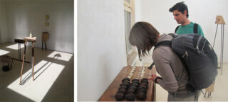 Zwei Personen untersuchen Objekte auf einem Tisch in einer minimalistischen, sonnendurchfluteten Galerie mit Holzskulpturen und Schatten auf dem Boden.