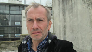 Ein Mann mittleren Alters mit kurzem, ergrauendem Haar steht im Freien neben einer Betonmauer, im Hintergrund sind moderne Bürogebäude zu sehen.