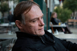 Ein Mann mittleren Alters mit kurzen Haaren sitzt im Freien an einem Cafétisch und blickt mit neutralem Gesichtsausdruck nach unten. Im Hintergrund sind unscharfe Personen und Sitzgelegenheiten im Freien zu sehen.