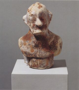 Büste eines älteren Mannes mit Schnurrbart, aus verwittertem und fleckigem Stein, auf einem schlichten Sockel vor grauem Hintergrund.