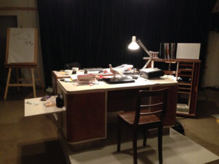 Vor einem dunklen Vorhang steht ein schwach beleuchteter Schreibtisch voller Papiere, Bücher und Büromaterial. Im Hintergrund stehen ein Holzstuhl, eine Schreibtischlampe und eine Staffelei.