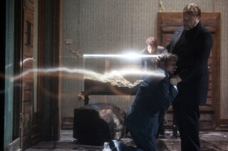 Ein Mann im Anzug kniet, während er von einem anderen Mann, der über ihm steht, gewürgt wird; eine dritte Person ist im Hintergrund und ein Lichtstrahl erstreckt sich über die Szene.