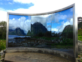 Eine Installation aus gebogenem Glas oder Spiegel im Freien spiegelt eine malerische Landschaft mit Bergen, Wasser, Felsen und Wolken unter einem teilweise bewölkten Himmel wider.