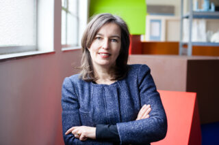 Eine Frau mit schulterlangem braunem Haar und einem blauen Blazer mit Strukturmuster steht mit verschränkten Armen in einem Gebäude, zu ihrer Linken ist ein Fenster, im Hintergrund sind geometrische Formen zu sehen.