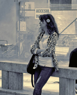 Eine Frau steht mit verschränkten Armen an einer Betonbarriere vor einem Zaun mit einem Schild mit der Aufschrift „ACCESUL INTERZIS“ gelehnt.