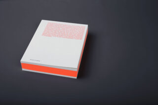 Ein dickes weißes Buch mit orangefarbenem Text auf dem Einband und einem leuchtend orangefarbenen Streifen entlang der Seitenränder, auf einer dunklen Oberfläche platziert.