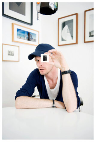 Ein Mann mit marineblauer Mütze und dunklem Pullover sitzt an einem Tisch und hält sich ein Dia vors Gesicht. An der Wand hinter ihm hängt ein gerahmtes Kunstwerk.