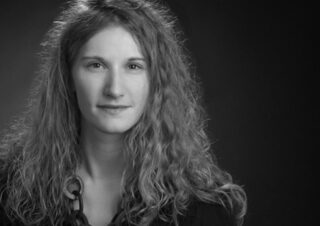 Schwarz-Weiß-Porträt einer Frau mit langen lockigen Haaren, die ein dunkles Oberteil und eine klobige Halskette trägt und mit neutralem Gesichtsausdruck vor einem schlichten Hintergrund in die Kamera blickt.