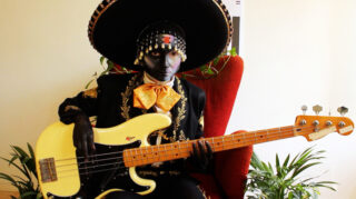 Person in Mariachi-Kleidung mit schwarz bemaltem Gesicht, sitzt auf einem roten Stuhl, hält eine gelbe E-Bassgitarre, umgeben von grünen Pflanzen.