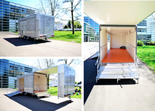 Drei Ansichten eines mobilen Anhängers mit Seitenrampen und offenem Innenraum, der an einem sonnigen Tag vor modernen Bürogebäuden geparkt ist.