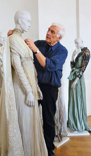 Ein älterer Mann in einem blauen Hemd richtet den Kragen eines detailreichen, hellen Kleides an einer Schaufensterpuppe, während im Hintergrund zwei weitere Schaufensterpuppen aufwendige Kleider tragen.