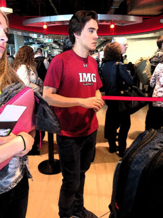 Ein junger Mann in einem T-Shirt der IMG Academy steht in einer geschäftigen Schlange im Innenbereich, umgeben von Menschen und Gepäck.
