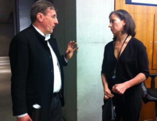 Ein Mann und eine Frau stehen im Haus und unterhalten sich. Der Mann gestikuliert mit der Hand, während die Frau mit einer Tasche in der Hand zuhört.