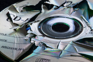 Eine Nahaufnahme einer CD-R-Disc, deren Oberfläche stark zerrissen und beschädigt ist, wodurch die darunterliegende reflektierende Schicht freigelegt wird.