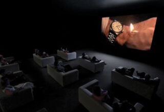 Eine Gruppe von Menschen sitzt auf Sofas in einem dunklen Raum und beobachtet einen großen Bildschirm, auf dem eine Nahaufnahme einer Armbanduhr zu sehen ist, die neben ein brennendes Streichholz gehalten wird.