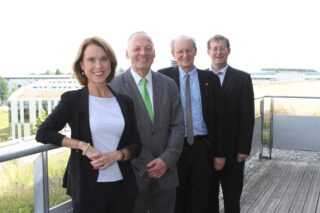 Vier Erwachsene in Geschäftskleidung stehen in einer Reihe im Freien auf einem Holzdeck und posieren für ein Gruppenfoto mit Gebäuden im Hintergrund.