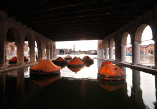 Aufblasbare orangefarbene Flöße treiben auf ruhigem Wasser unter einem großen Steingebäude mit Bögen und freiem Blick auf einen entfernten Glockenturm bei Sonnenuntergang.