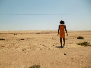 Eine Person in einem orangefarbenen Kleid geht allein unter klarem Himmel durch eine flache, trockene Wüstenlandschaft.
