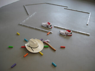 Ein weißer Cowboyhut, eine Spielzeugaxt, bunte Kreidestifte und ein Paar Turnschuhe sind auf einem grauen Boden neben Umrissen aus durchscheinenden Balken angeordnet.