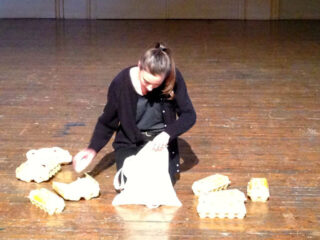 Eine Person sitzt auf einem Holzboden und legt Eierkartons in eine weiße Tüte. Mehrere Kartons liegen verstreut herum.