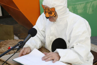 Eine Person in einem weißen Kostüm mit Tigermaske liest vor einem Mikrofon aus einem offenen Buch.