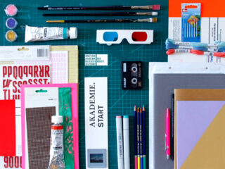 Verschiedene Kunst- und Büroartikel, darunter Farben, Pinsel, Stifte, Bleistifte, farbiges Papier, Faden, Kassetten, 3D-Brillen und Schneidewerkzeuge, angeordnet auf einer grünen Schneidematte.