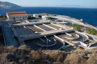 Eine Kläranlage an der Küste mit mehreren Freilufttanks, Gebäuden und umgebender trockener Vegetation mit Blick auf das Meer und die Hügel in der Ferne.
