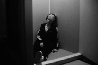 Eine Person mit Kopfhörern und einem Adidas-Trainingsanzug sitzt mit geschlossenen Augen auf dem Boden in einer schwach beleuchteten Ecke und lehnt an der Wand.