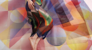 Eine Person sitzt barfuß auf einer Oberfläche, die mit buntem, durchscheinendem Stoff mit geometrischem Muster bedeckt ist, und trägt ein transparentes Kleid mit ähnlichem Muster.