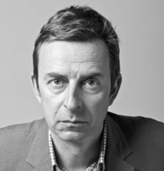 Ein Mann mit kurzen Haaren, einem Jackett und einem karierten Hemd, starrt mit neutralem Gesichtsausdruck vor einem schlichten Hintergrund direkt in die Kamera.