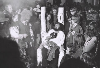 Ein bärtiger Mann sitzt auf einem großen, weißen Holzstuhl, umgeben von einer Menschenmenge, die ihn in einem Innenraum beobachtet.