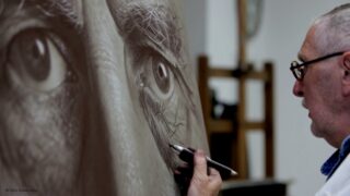 Ein Künstler skizziert ein detailliertes, realistisches Auge auf einer großen Leinwand. Dabei hält er einen Bleistift in die Nähe des Papiers, im Hintergrund ist eine Staffelei zu sehen.