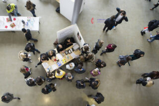 Draufsicht auf Menschen, die sich bei einer Veranstaltung in geschlossenen Räumen um einen Informationstisch versammelt haben, während andere in der Nähe auf einem polierten Betonboden gehen oder stehen.