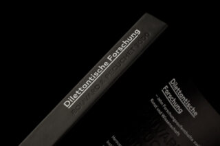 Nahaufnahme des Buchrückens und eines Teildeckels eines schwarzen Buches mit dem Titel „Dilettantische Forschung“ von Bouvard & Pecuchet 3000 mit weißer Schrift.