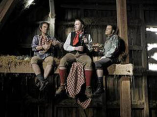 Drei Männer in traditioneller bayerischer Kleidung sitzen auf einem Heuboden in einer rustikalen Holzscheune, über dessen Rand ein kariertes Tuch hängt.