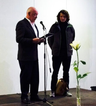 Ein älterer Mann spricht mit einem Ordner in ein Mikrofon, während eine jüngere Person neben ihm auf der Bühne steht; im Vordergrund ist eine einzelne weiße Blume in einer Vase zu sehen.