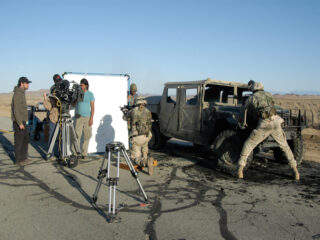 Filmteam und Schauspieler in Militäruniformen auf einer Wüstenstraße, mit einem ausgebrannten Fahrzeug, Kameraausrüstung und Lichtvorhang im Blick.