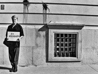 Eine Person steht an einer Wand und hält ein Schild mit der Aufschrift „Zu Verschenken“. Die Wand dahinter ist fleckig und hat ein vergittertes Fenster.