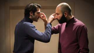 Zwei Männer stehen sich in einem Raum gegenüber. Einer zeigt mit dem Finger auf die Stirn des anderen, während der andere die Stirn des ersten Mannes berührt. Beide wirken ernst.